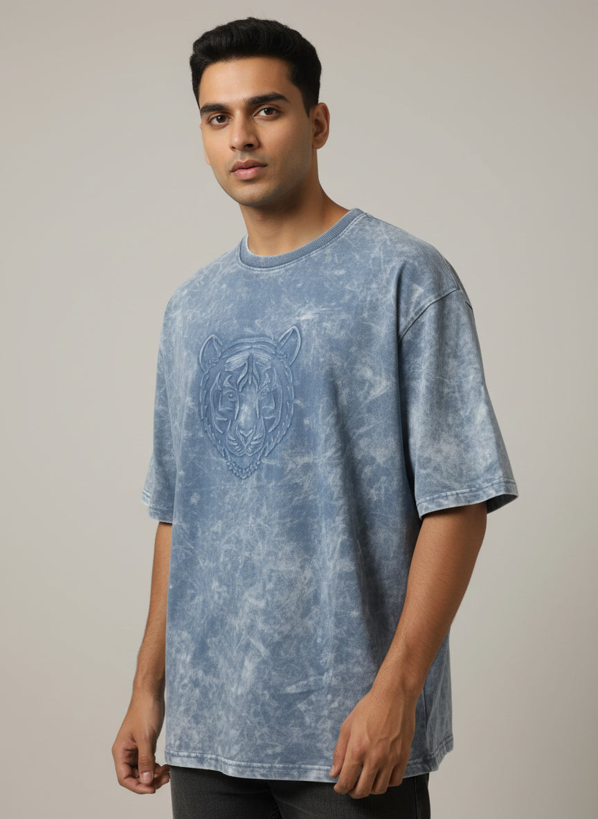Blue acid-wash t-shirt with a tiger design on a hanger against a beige wall.